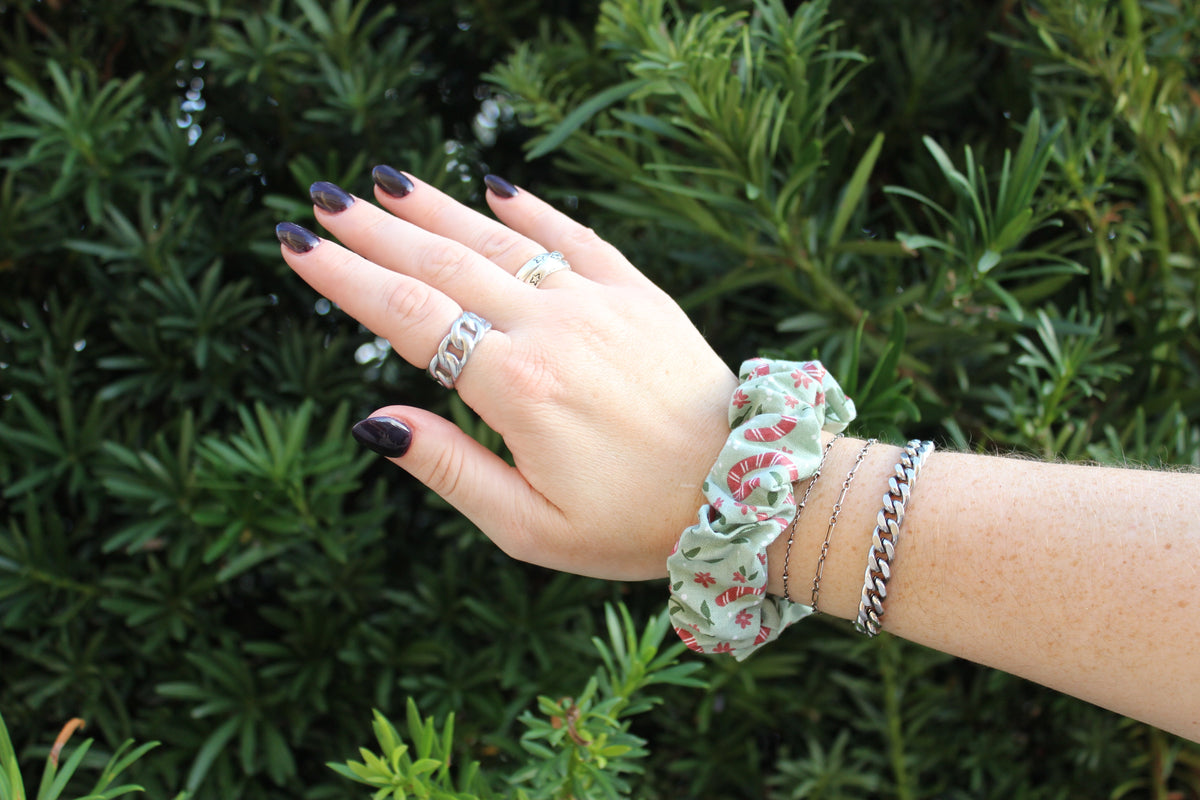 Hand wearing a green and red candy cane patterned hair scrunchie and silver rings against a leafy background.