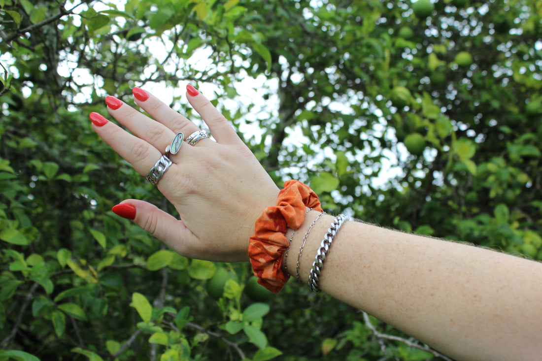 Hand wearing an orange ruffled hair scrunchie, silver rings, and red nail polish against a leafy green background.