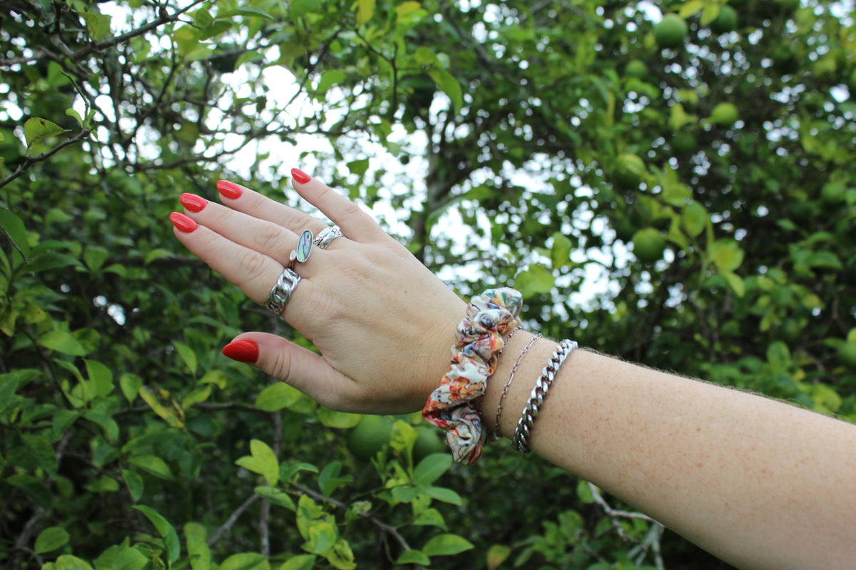 Hand wearing silver rings and a colorful patterned fabric hair scrunchie against a backdrop of green leaves.