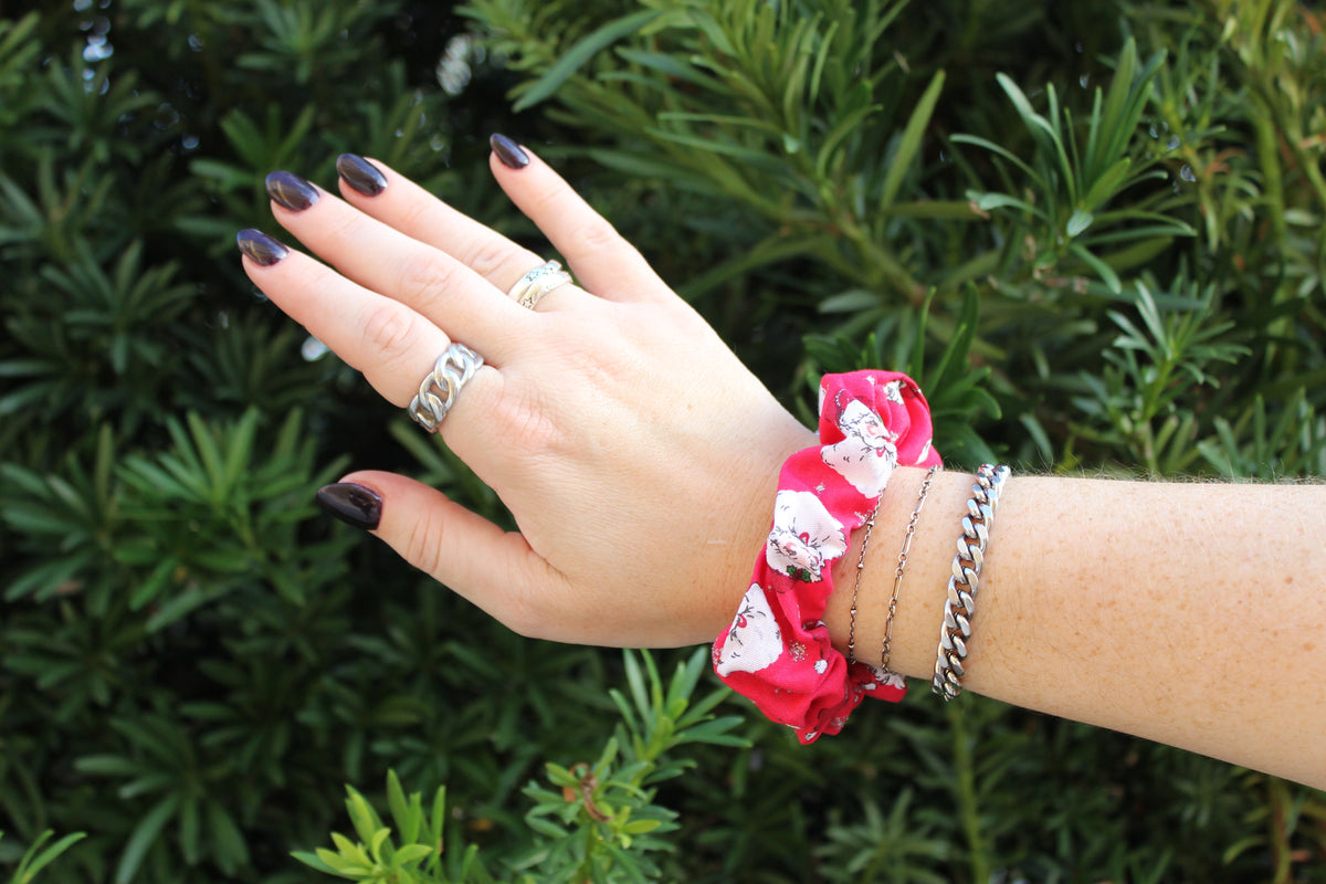 Red and white floral hair scrunchie worn on a wrist beside silver chain bracelets against a green leafy background.