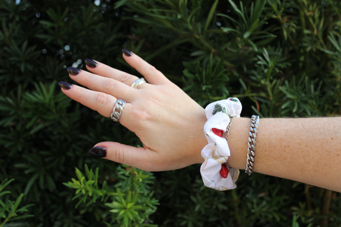 White ruffled scrunchie with red berry details worn on a wrist next to a silver chain bracelet.